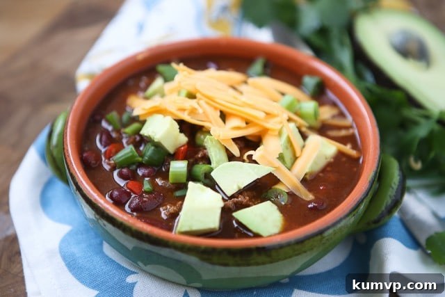 A close-up of the delicious Chipotle Chicken Chili, garnished generously with vibrant shredded cheese, freshly diced avocado, and crisp green scallions, showcasing its irresistible texture and color.