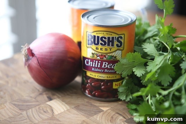 An inviting scene featuring a rustic wooden cutting board adorned with a vibrant red onion, two cans of Bush's Chili Kidney Beans, and a fresh bunch of cilantro, highlighting key ingredients for the Chipotle Chicken Chili recipe.