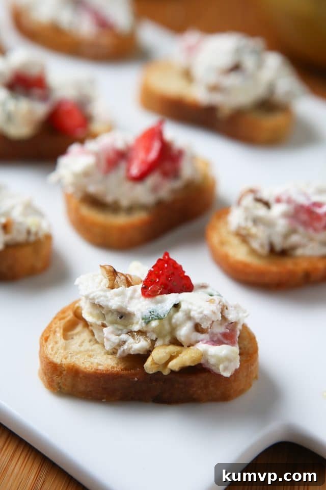 Goat Cheese Spread with Strawberries and Basil A single piece of toasted baguette topped with a generous dollop of Strawberry Basil Goat Cheese Spread, garnished with fresh strawberry pieces, basil, and walnuts, showcasing its perfect texture and vibrant appeal on a pristine white platter.
