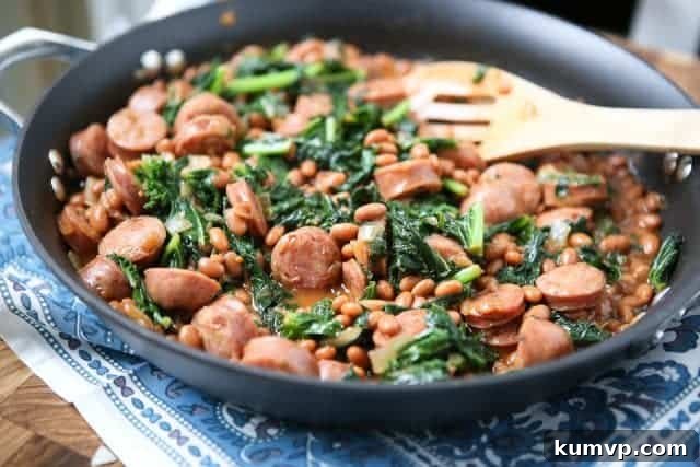 Baked Bean, Sausage and Kale Skillet - A Delicious One-Pan Meal skillet with chicken sausage, baked beans, and kale with wooden cooking spoon resting on it