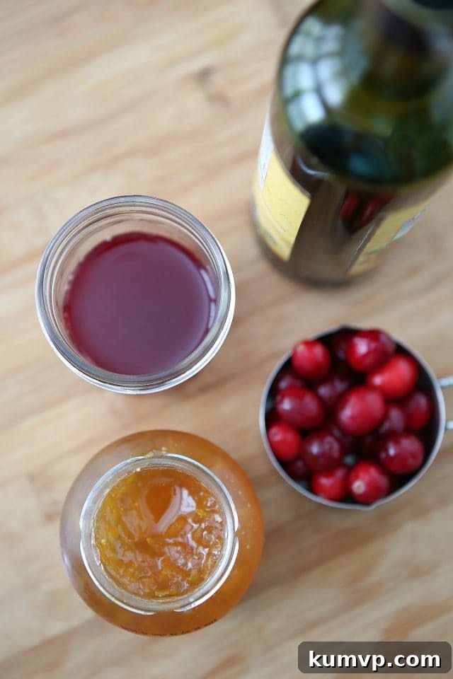 Close-up shot of key ingredients for Cranberry Balsamic Chicken Skillet: a jar of orange marmalade, fresh cranberries in a container, and a glass of 100% unsweetened cranberry juice with its bottle, all arranged on a rustic wooden countertop, ready for cooking.
