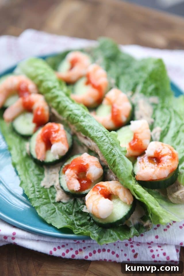 Two crisp romaine lettuce leaves generously stuffed with succulent sriracha shrimp, creamy hummus, and refreshing cucumber slices, artfully drizzled with a vibrant red hot sauce, served on a pristine white plate – a perfect low-carb, high-protein meal for weight loss