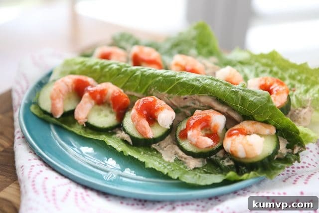 A plate featuring two beautifully prepared romaine lettuce wraps, generously filled with spicy Sriracha shrimp, creamy hummus, and crisp cucumber, garnished with a vibrant drizzle of hot sauce, ready to be enjoyed as a light and healthy meal