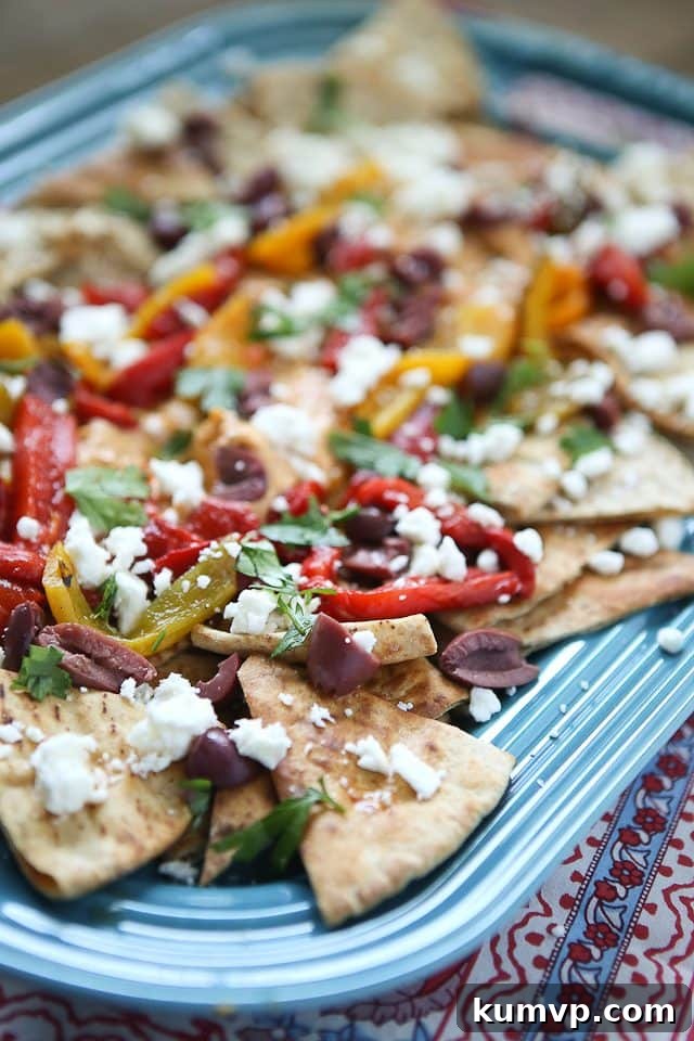A vibrant platter of Mediterranean Nachos, featuring golden pita chips generously topped with creamy Sabra Roasted Red Pepper Hummus, colorful roasted peppers, briny Kalamata olives, crumbled feta cheese, and a sprinkle of fresh herbs.