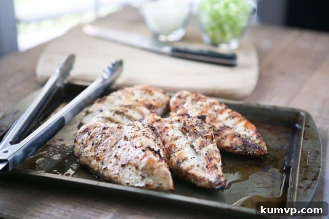 Four perfectly grilled lemon pepper chicken breasts cooling on a baking sheet, with tongs resting beside them, ready for dicing.