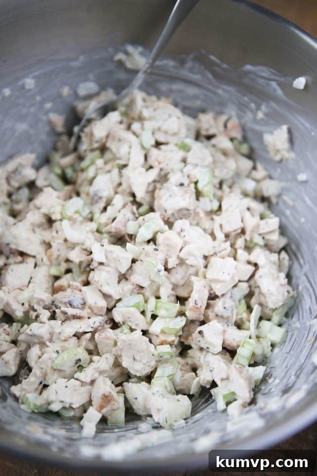 A close-up view of a mixing bowl where diced chicken, sweet onions, celery, and seasonings are being thoroughly combined with the creamy Greek yogurt and mayonnaise dressing.