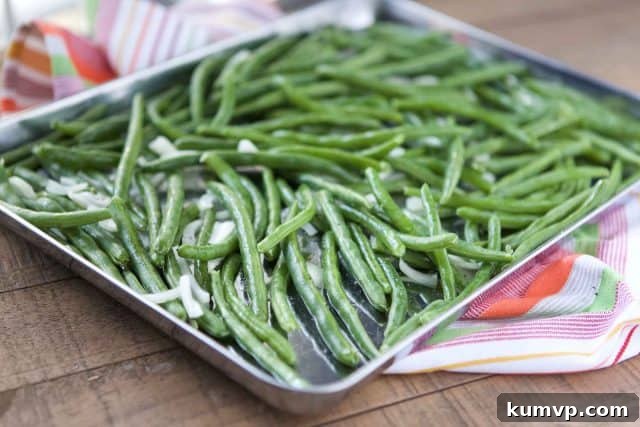 A sturdy baking sheet filled with an even layer of fresh, vibrant green beans and thinly sliced shallots, lightly coated with olive oil and seasoned, poised for roasting in the oven.