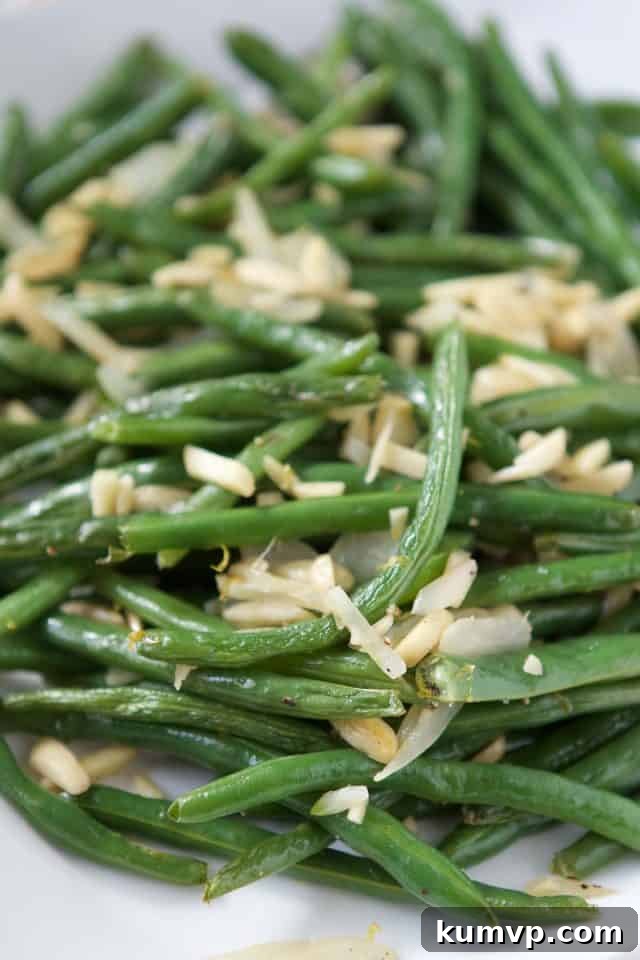 An extremely detailed close-up shot of roasted green beans, showcasing their tender-crisp texture, perfectly golden-brown slivered almonds, and tiny, vibrant flecks of lemon zest. The image highlights the beautiful browning and seasoning.