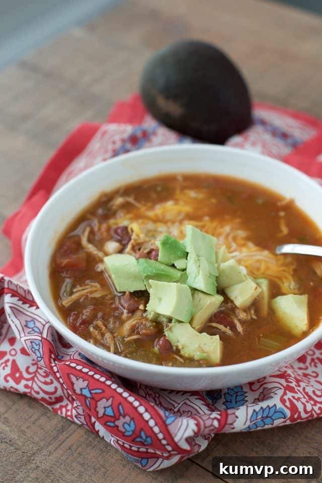 Instant Pot Mexican Chicken Soup A beautifully presented white bowl of Instant Pot Mexican Chicken Soup, garnished with fresh diced avocado, resting on a vibrant red and blue patterned napkin. A whole avocado in the background suggests freshness and flavor.