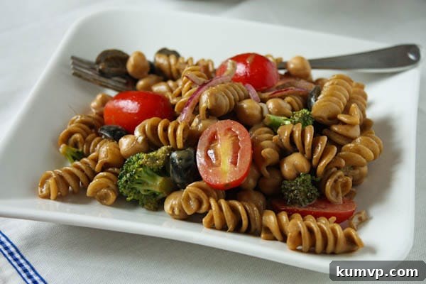 A close-up of a serving of pasta salad with cherry tomatoes, olives, red onions, and broccoli on a white plate with a fork